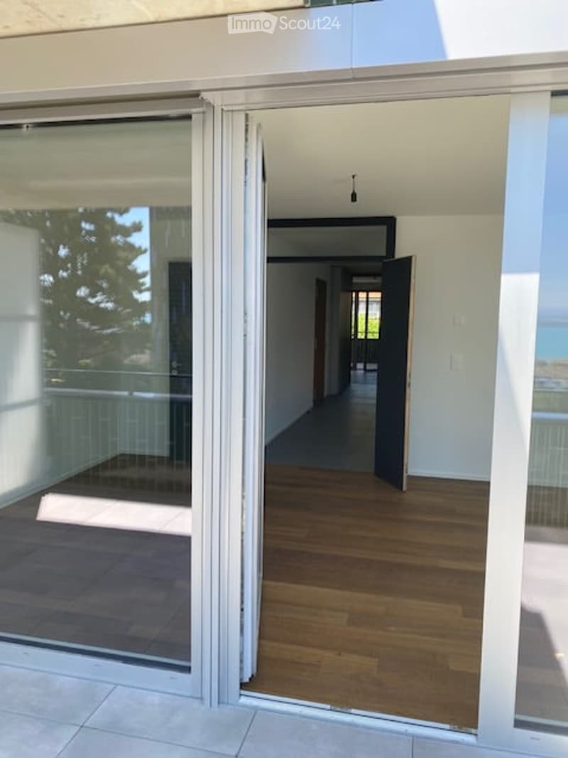 Modern hallway with wooden flooring, white walls, and large glass sliding doors leading to a balcony area.