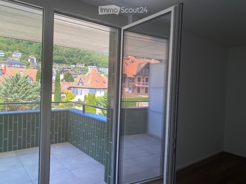Balcony with tiled floor, green railing, and open sliding doors leading to a room with a view of a town and greenery.