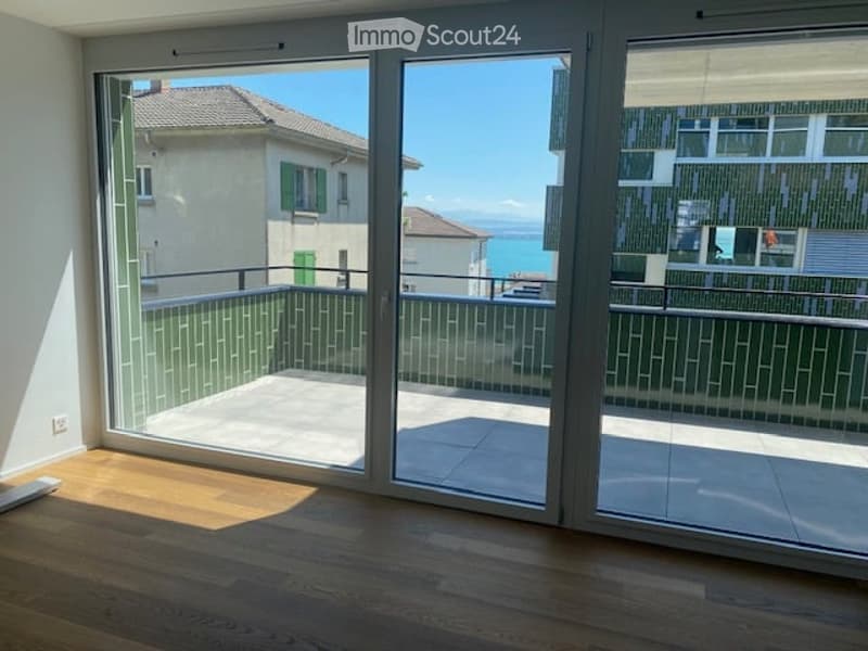 Modern balcony with large glass sliding doors, wooden floor, and a view of the sea and surrounding buildings.