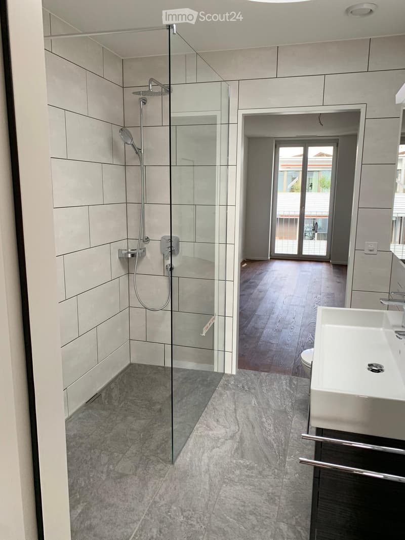 Modern bathroom with a glass-enclosed shower, a white sink, and a toilet. The floor is tiled in grey marble, and the walls are tiled in light grey.