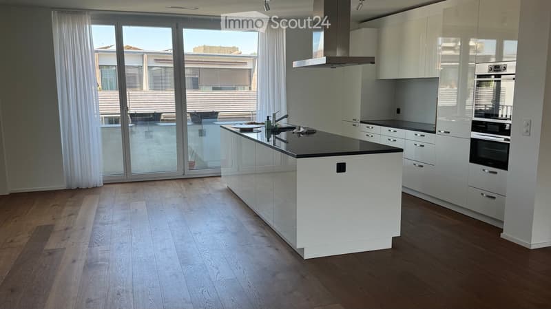 Modern kitchen with white cabinets, a central island, and a large window overlooking a balcony. Features include a stainless steel oven, a range hood, and a sleek black countertop.