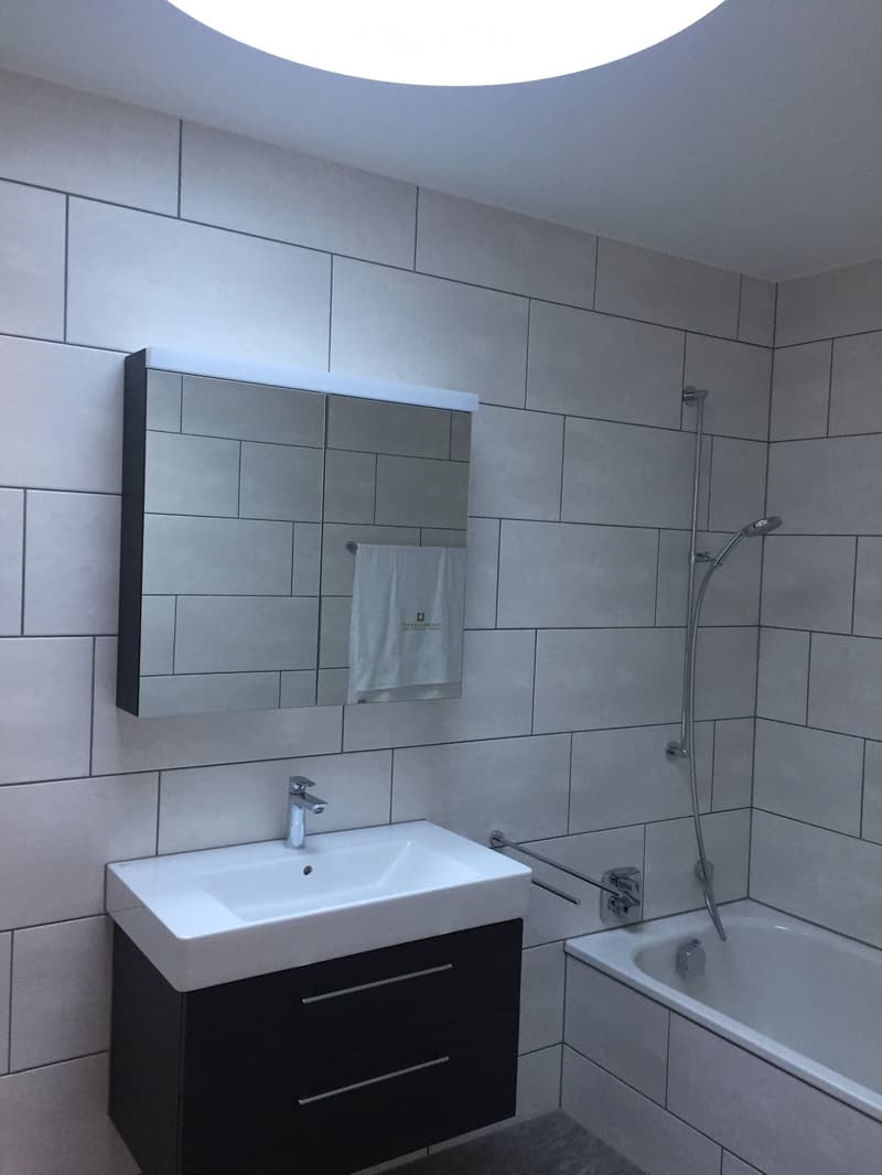 Modern bathroom with white tiles, a white sink with a chrome faucet, a dark wooden cabinet, a large mirror, a towel, and a bathtub with a showerhead.