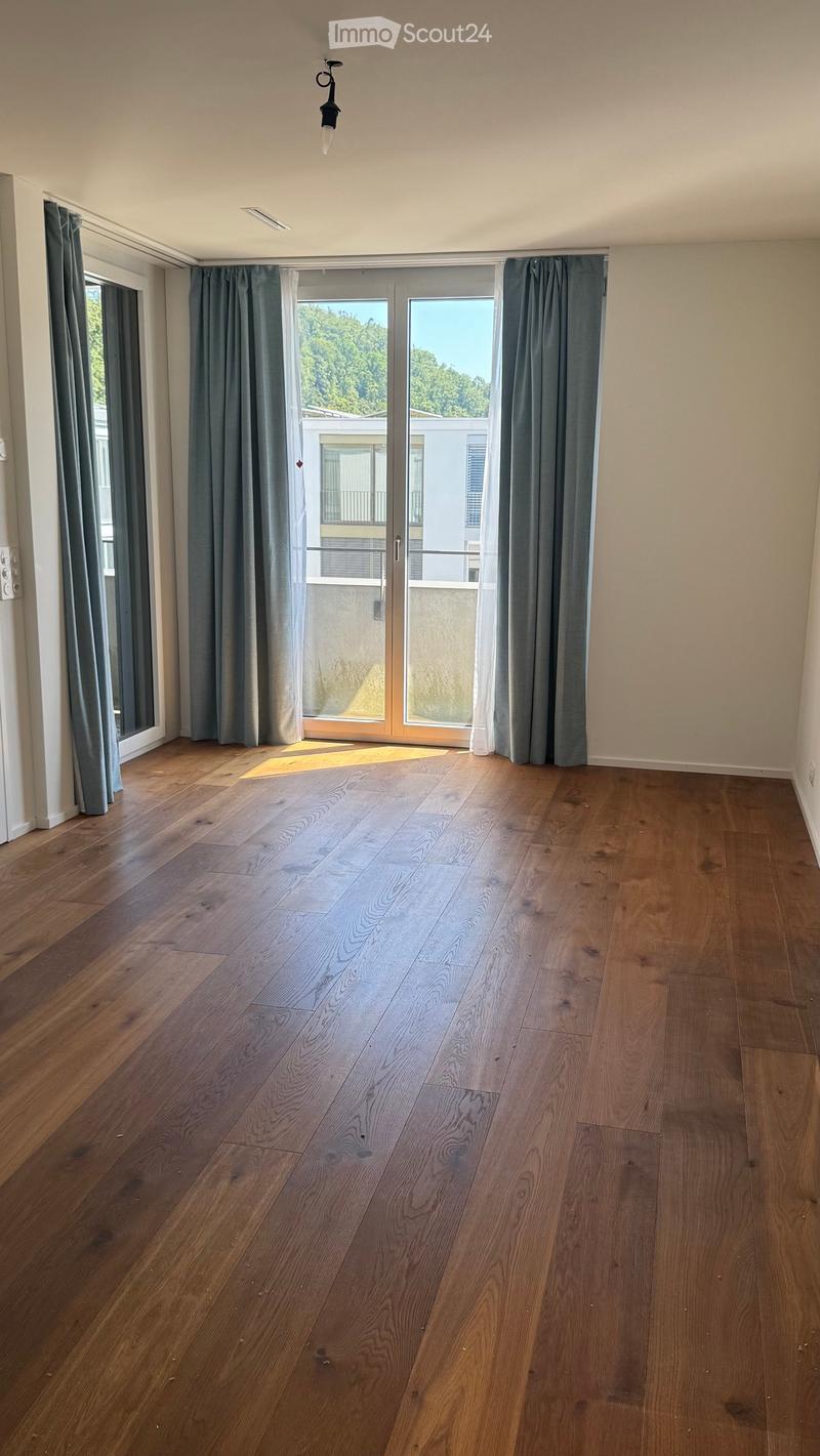 Empty room with wooden flooring, large windows with grey curtains, and a sliding glass door leading to a balcony.