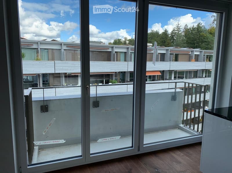 Balcony with glass doors opening to a modern residential complex, featuring white brick walls, railings, and a clear blue sky with scattered clouds.