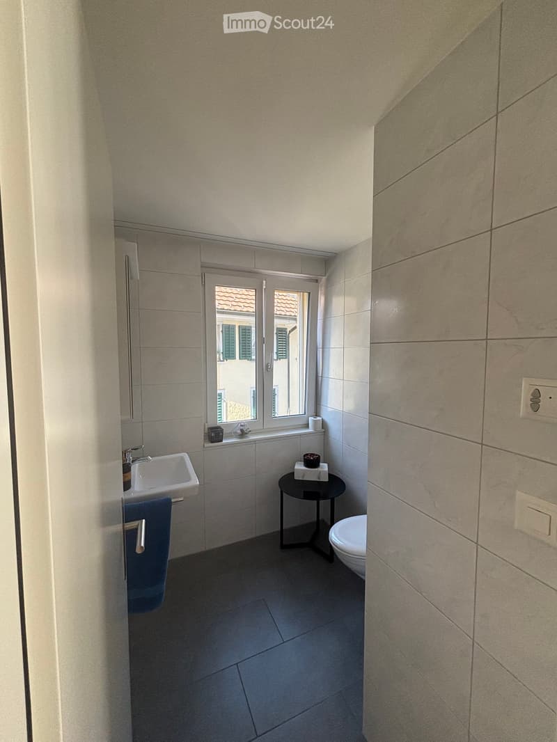 Modern bathroom with a sink, toilet, small black stool, and a towel hanging on the wall. The room has tiled walls and a window offering a view of a tiled roof.