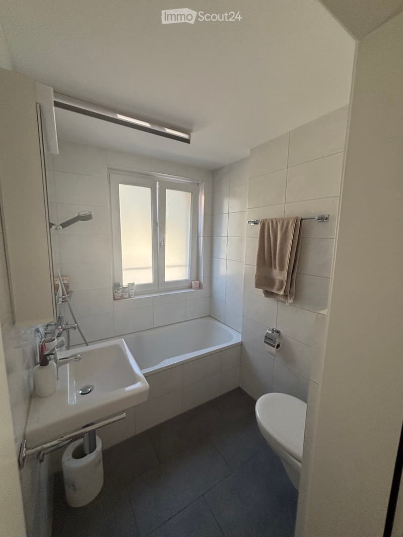 Modern bathroom with white tiles, a bathtub, sink, toilet, mirror, and a towel hanging on a rack. A window provides natural light.