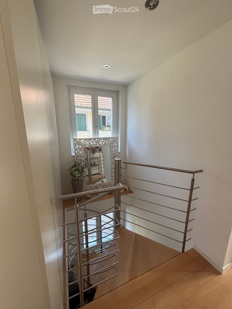 Modern hallway with wooden flooring, metal railing, and a decorative mirror on the wall. A vase with flowers is placed near the mirror.