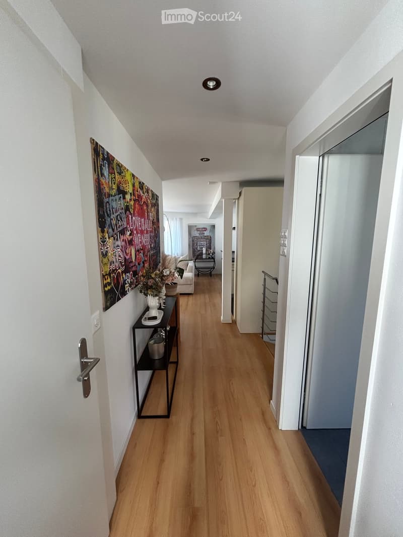 A long, well-lit hallway with wooden flooring, white walls, and modern decor including a small black shelf with decorative items and a colorful wall art piece.