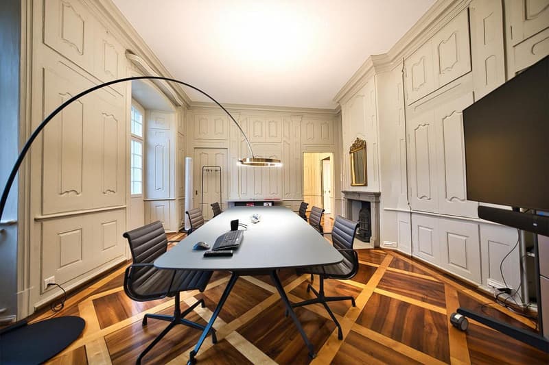 Elegant conference room with a long table, chairs, a keyboard, mouse, and a large screen. The room features ornate wooden paneling, a fireplace, and a large arched window.