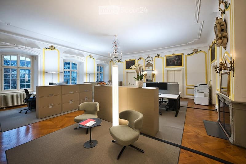 Elegant office space with wooden floors, large windows, and ornate mirrors. Features include a reception area with a cabinet, a seating area with two chairs and a small table, and a workspace with a desk and computer screen.