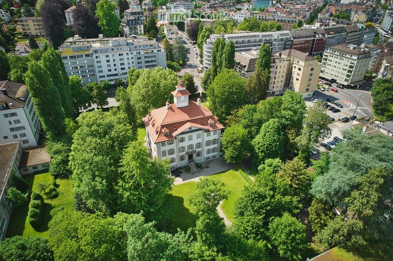 Aerial view of a large, classical-style building with a red-tiled roof and white facade, surrounded by lush green trees and a well-maintained lawn, situated in an urban area with other buildings and roads visible in the background.