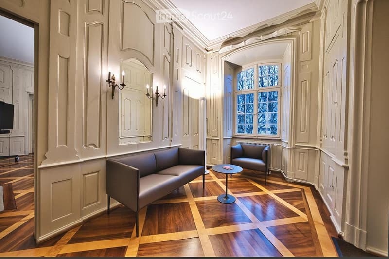 Elegant living room with ornate white paneling, hardwood floor, grey sofa, armchair, small round table, wall sconces, and a large mirror.
