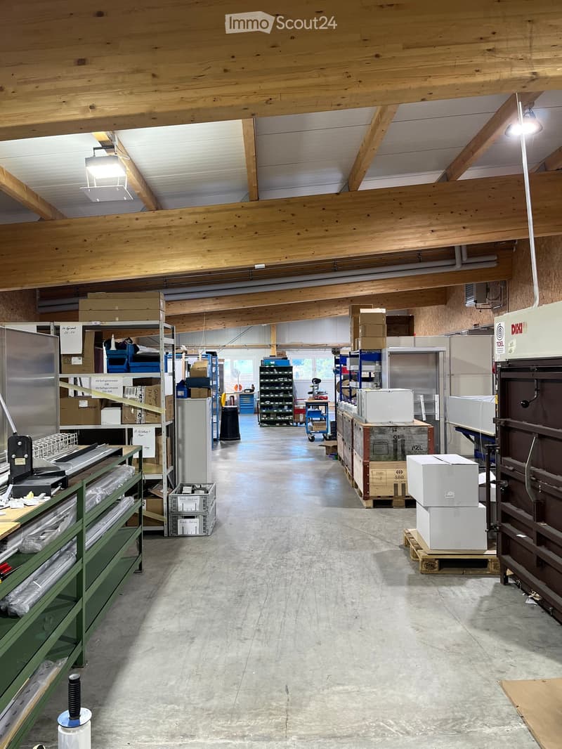 A spacious industrial or storage area with exposed wooden beams, concrete floor, and various storage equipment including shelves, refrigerators, and stacked cardboard boxes.