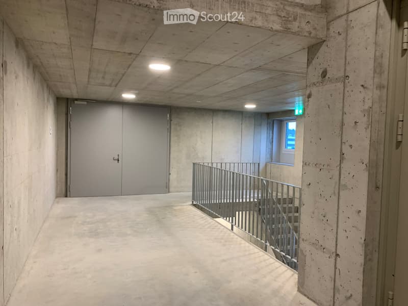 Concrete corridor with a staircase leading upwards, featuring a metal railing and a closed door at the end.