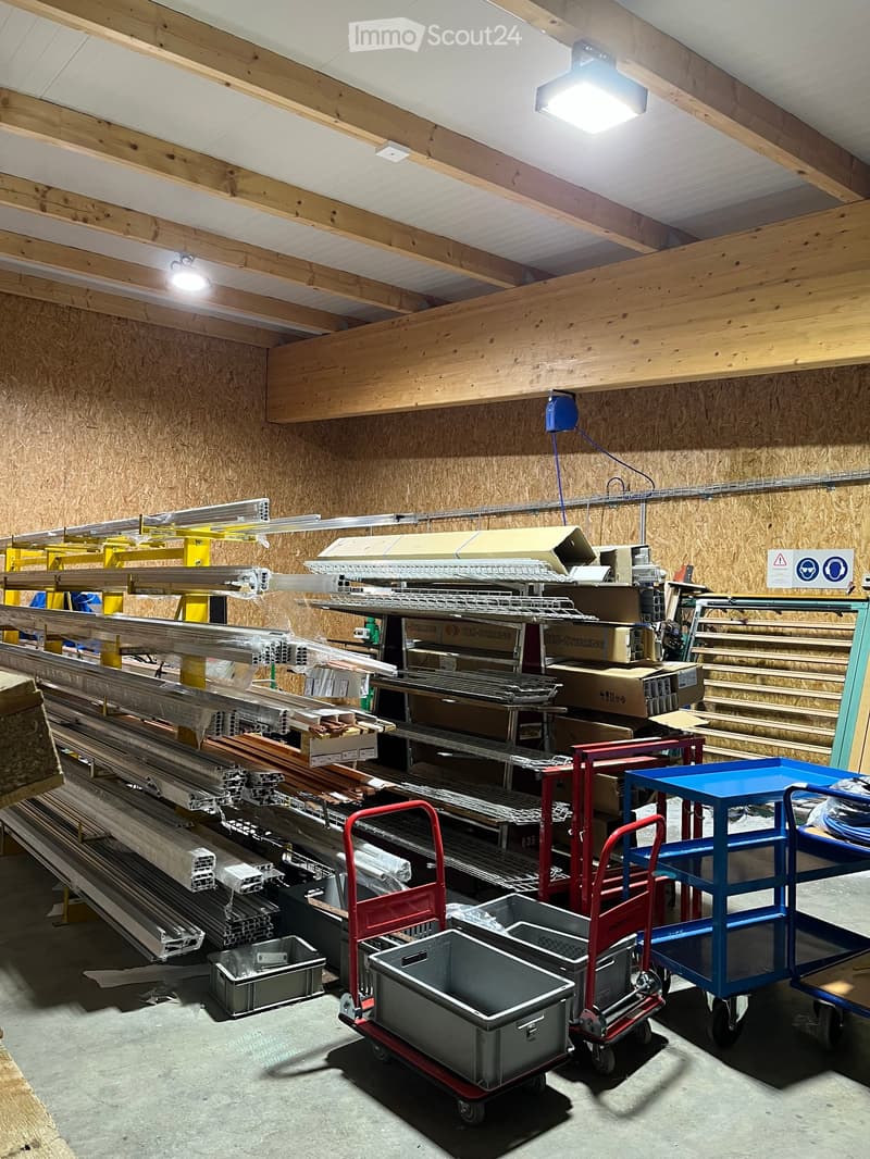 A storage area with exposed wooden beams and walls, filled with metal bars, boxes, and carts. The space is well-lit with overhead lights and has a concrete floor.