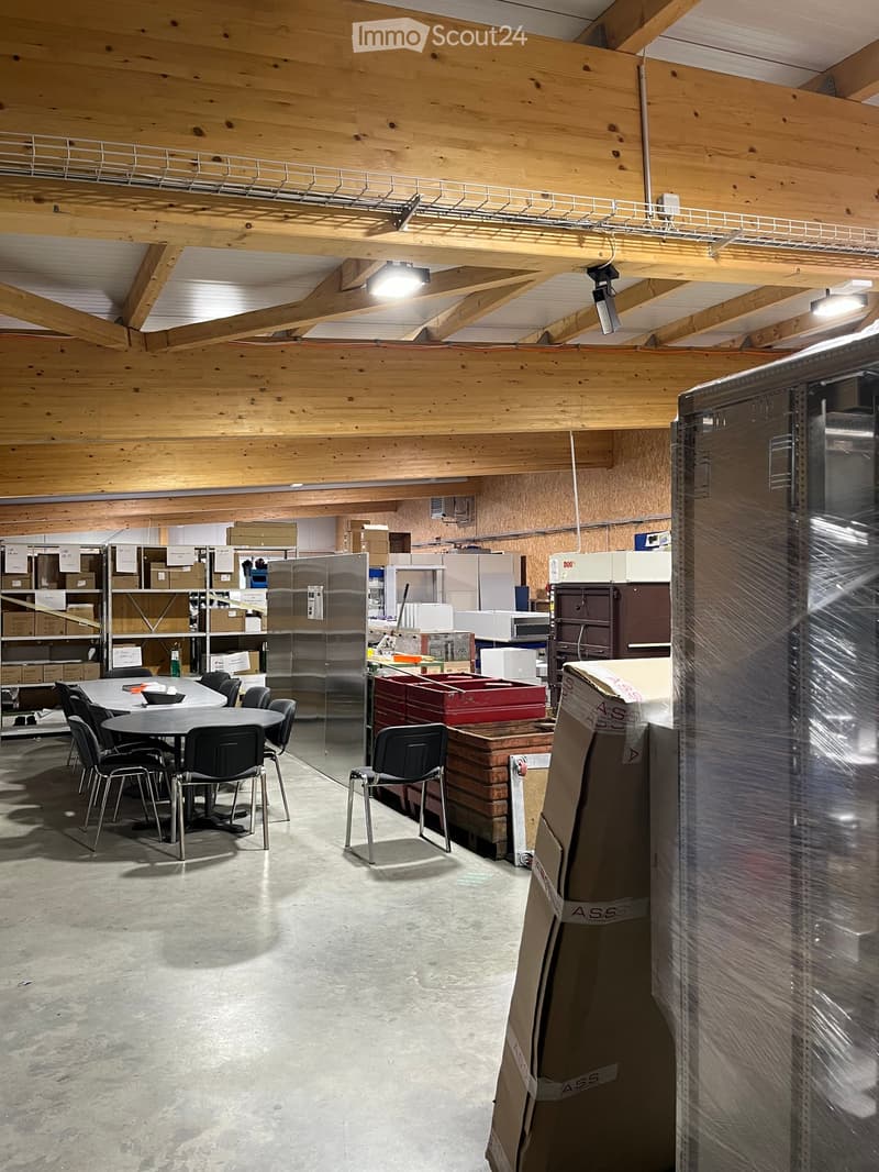 A spacious, unfinished room with exposed wooden beams and a concrete floor. The room features a round table with chairs, a refrigerator, microwave, oven, shelves with boxes, and various storage items.