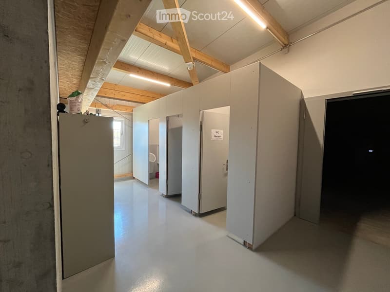 Empty hallway with white walls, grey floor, and several stall doors leading to bathrooms.
