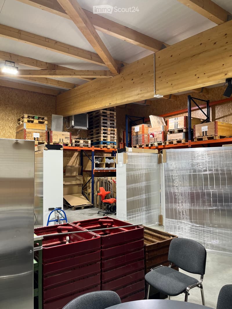 A spacious, unfinished room with exposed wooden beams and plywood walls, featuring various storage items including shelves, pallets, and storage boxes. There are a few chairs and a table in the foreground.