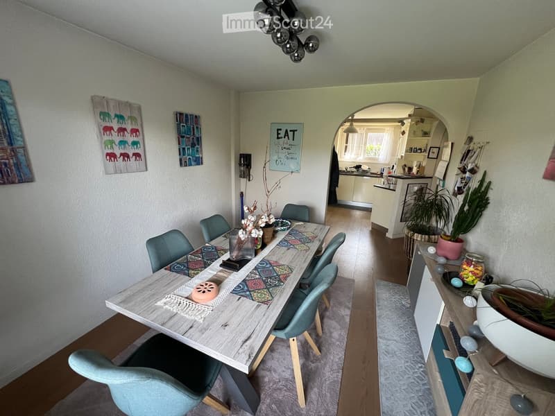 Dining area with a wooden table, teal chairs, and decorative items. Open-plan kitchen visible in the background with cabinets, sink, and appliances.