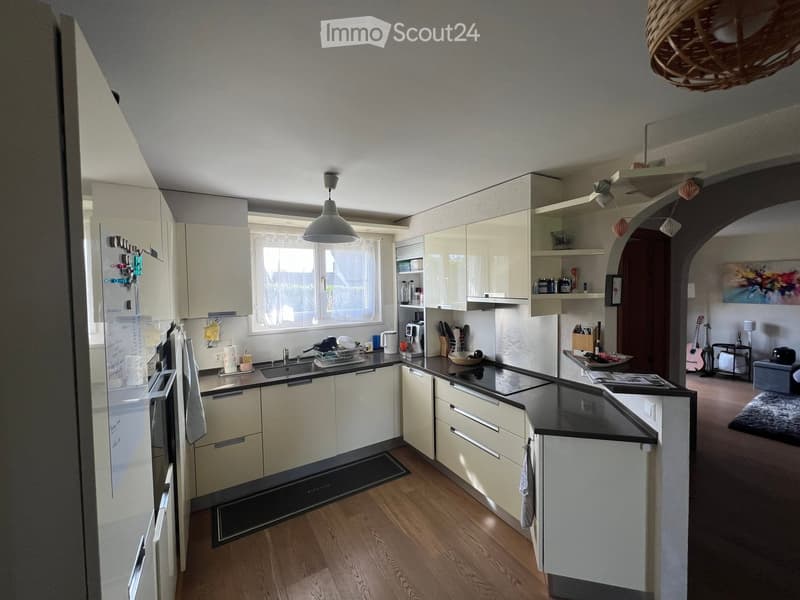 Modern kitchen with light-colored cabinets, black countertops, stainless steel appliances, and a central island. Features a window, pendant lights, and a small living area with a guitar and painting visible in the background.