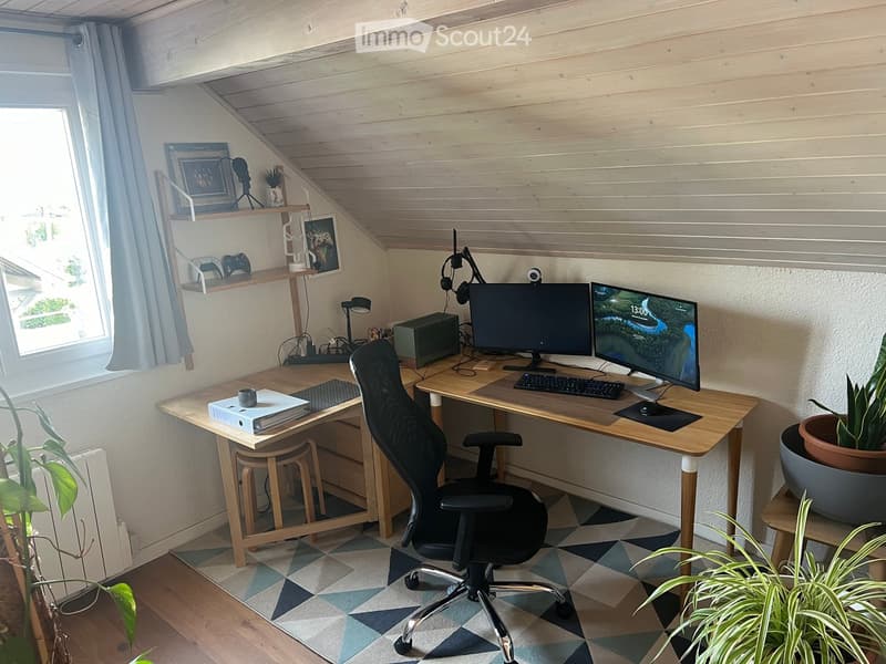 Cozy attic workspace with wooden desks, ergonomic chair, dual monitors, and decorative plants.