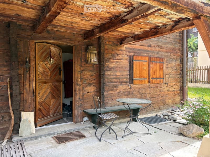 Wooden house exterior with a covered porch area featuring a table, chairs, and a bench, surrounded by stone and greenery.