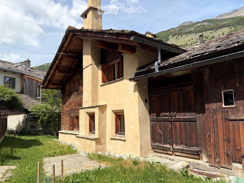 Two-story rustic house with wooden shutters, stone foundation, and a chimney. The house is surrounded by a grassy area and is set against a mountainous backdrop.
