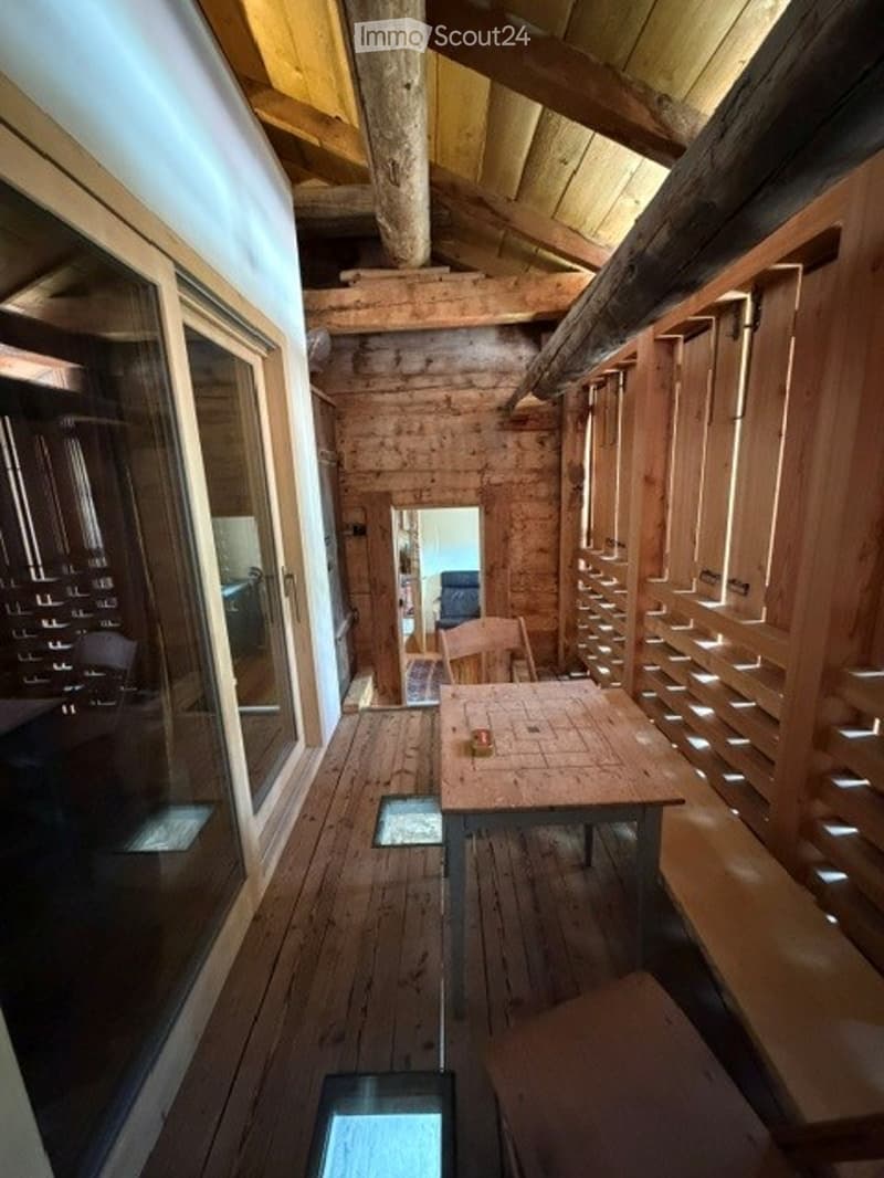 A rustic wooden room with exposed beams and plank walls, featuring a wooden table and chair, and large windows with wooden shutters.