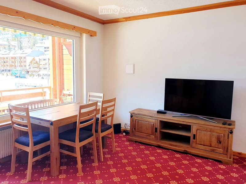 A furnished room with a wooden dining table, four chairs, a television on a wooden stand, and a large window with a view of the outside area.