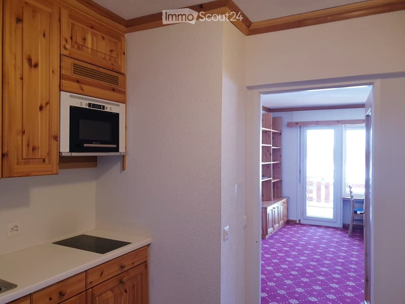 The image shows a small kitchen with wooden cabinets, an oven, and an induction stove. The kitchen has a white countertop and a pink floral-patterned floor. An open doorway leads to another room with shelves and a chair visible.