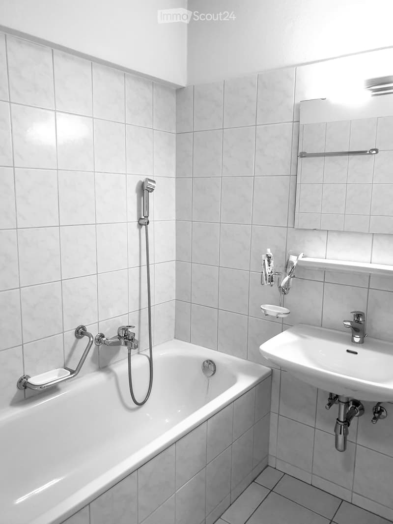 Modern bathroom with white tiled walls and floor, featuring a bathtub with a handheld showerhead, a sink with a modern faucet, and a mirror above the sink.