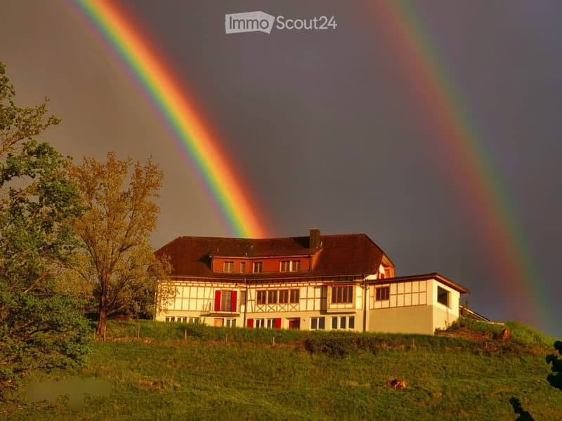 Hilltop house, two rainbows, brown roof, red shutters, white and brown walls, surrounded by grass and trees
