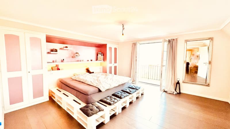 A bedroom with a wooden floor, pink and white walls, a bed made with white wooden pallets, a balcony entrance with railings, a mirror, a cabinet with pink doors, a shelf with various items, and a lamp.