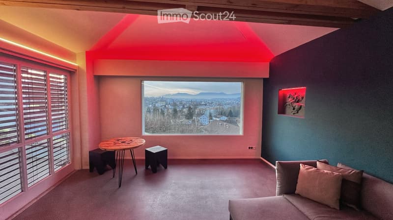 Pink and blue walls, large window with city view, round wooden table with metal legs, two stools, couch with pillows, decorative light on wall
