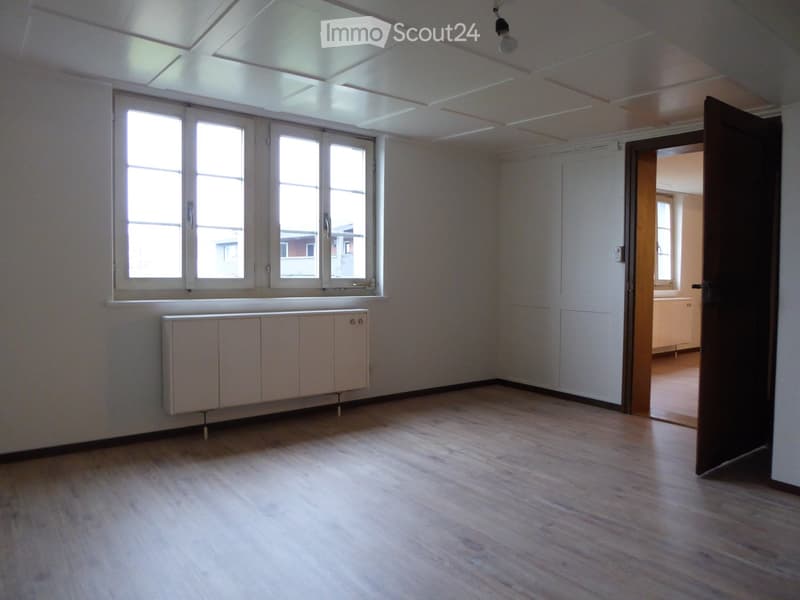 Empty room with wooden flooring, white walls, large windows, and a radiator. An open doorway leads to another room.