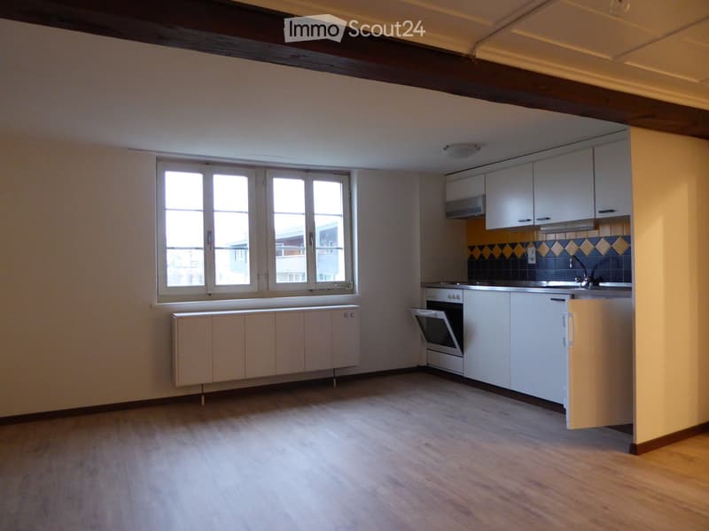 Empty room with wooden flooring, white cabinets, and a window with multiple panes. The room includes a radiator and a small kitchen area with built-in appliances.
