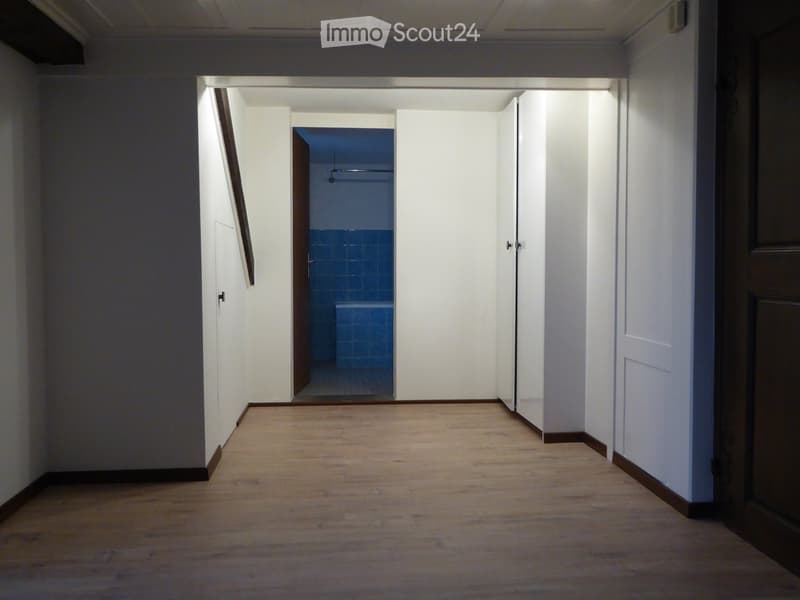 Empty hallway with wooden flooring, white walls, and doors leading to other rooms, including a bathroom with blue tiles visible at the end.