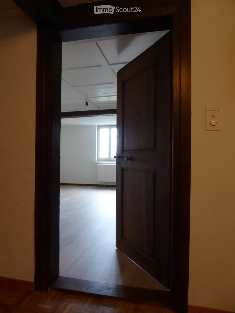 Empty room with wooden flooring and white walls, visible through an open wooden door.