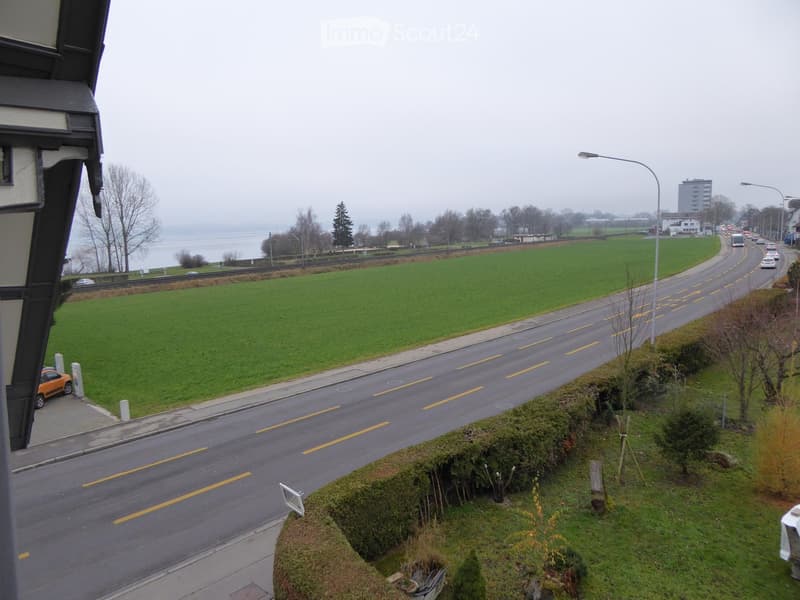 Balcony view showing a road with traffic, a green field, and a distant body of water under an overcast sky.