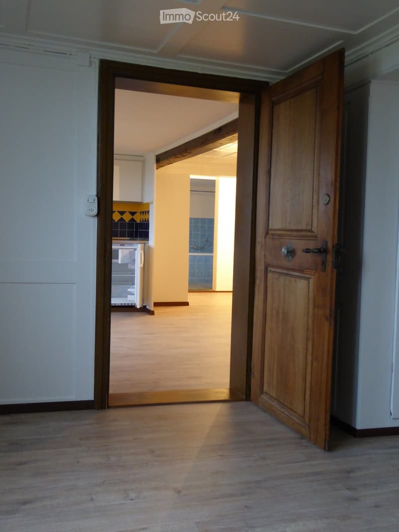 Empty hallway with wooden flooring and white walls, leading to an open door that reveals a brightly lit room with a visible kitchen area in the background.