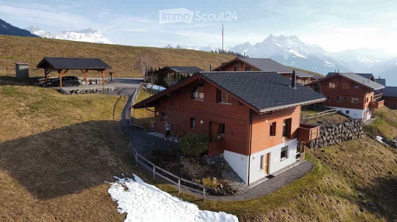Wooden chalet, black roof, white walls, surrounded by snow, mountains in the background, car parked in front