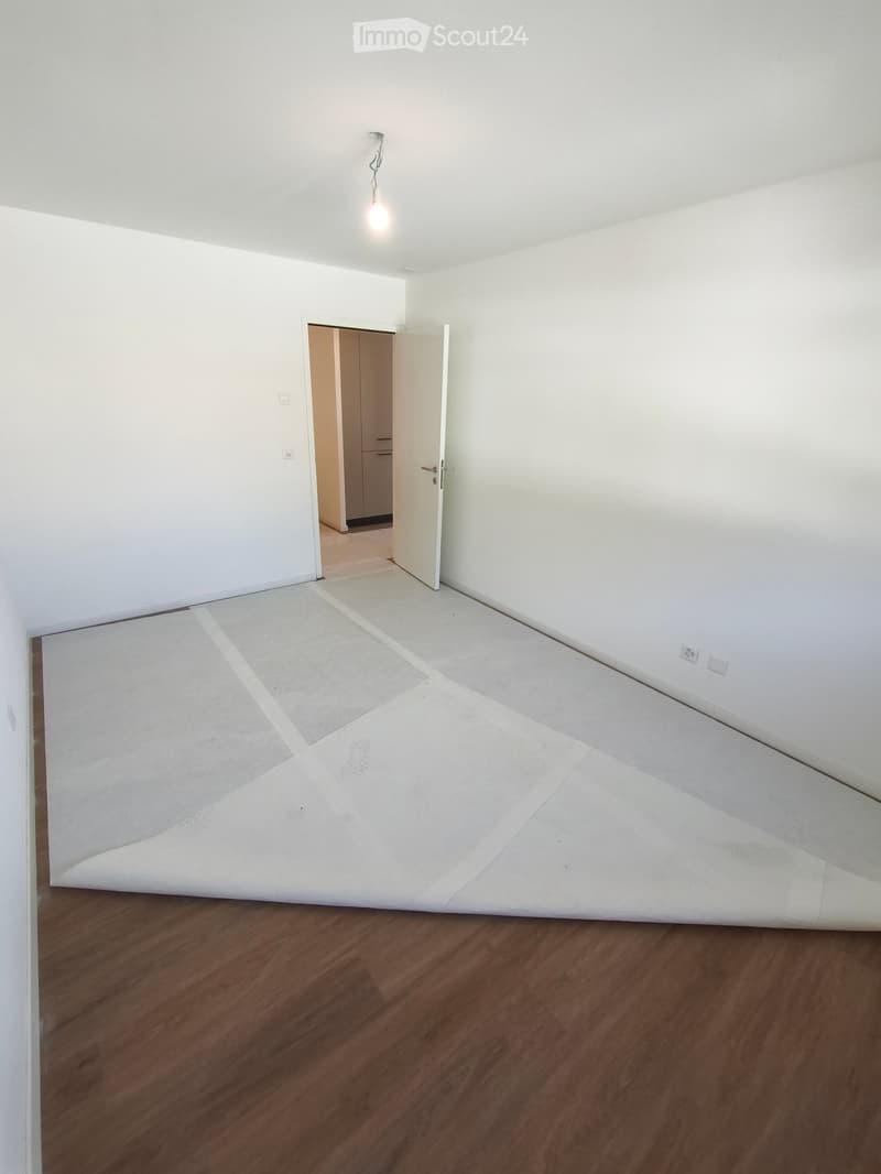 Empty room with wooden flooring, white walls, and a partially laid carpet. A doorway leads to another room with light-colored walls and a closet.