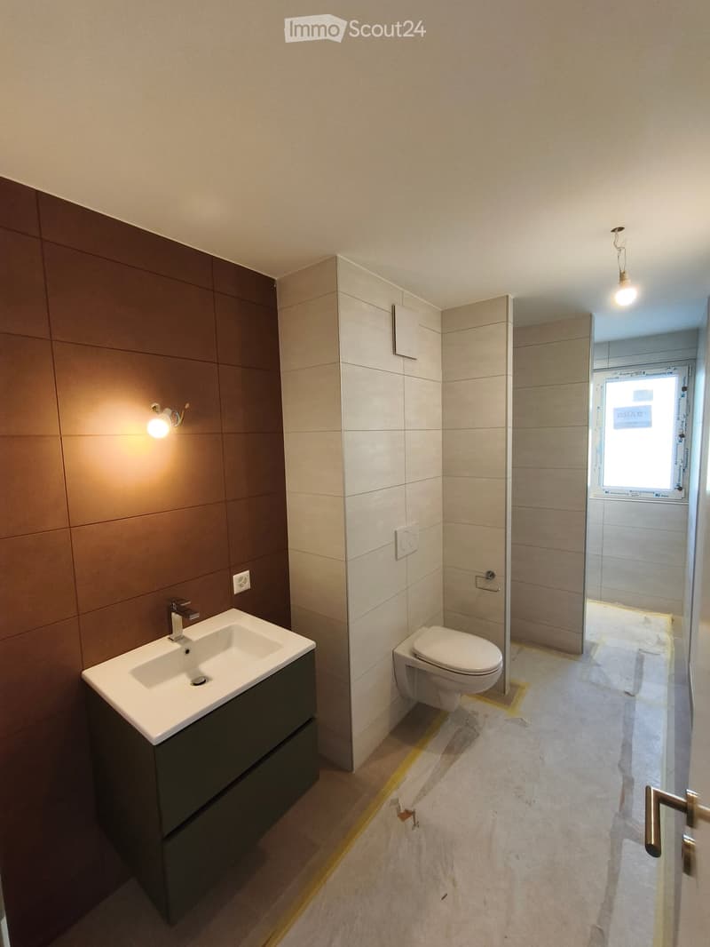 Modern bathroom with a white sink, dark green cabinet, toilet, and a glass shower enclosure. Light-colored tiles and warm lighting.