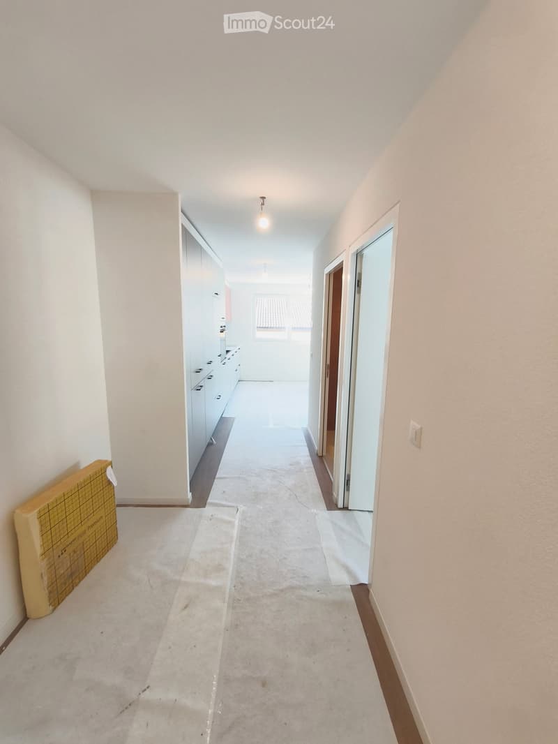 Empty hallway with white walls, carpeted floor, and recessed lighting. Doors lead to adjacent rooms, and a large cardboard box is placed against the left wall.