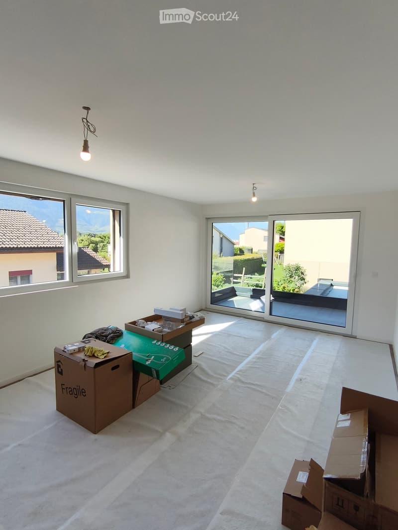 Empty room with white walls, large windows, and sliding glass doors leading to a balcony. Several cardboard boxes are placed around the room.