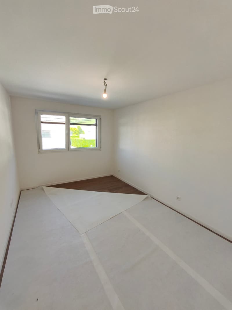 Empty room with white walls, wooden flooring, and a large window letting in natural light. A light fixture hangs from the ceiling.
