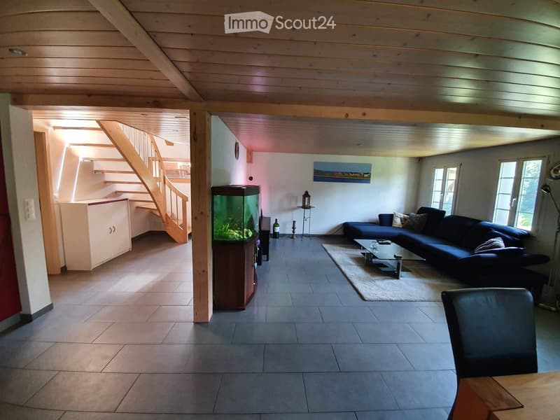 A spacious living room with a high wooden ceiling, large windows, a staircase leading to an upper level, a blue sectional sofa, a glass coffee table, a fish tank, and a wooden dining table with a black leather chair.