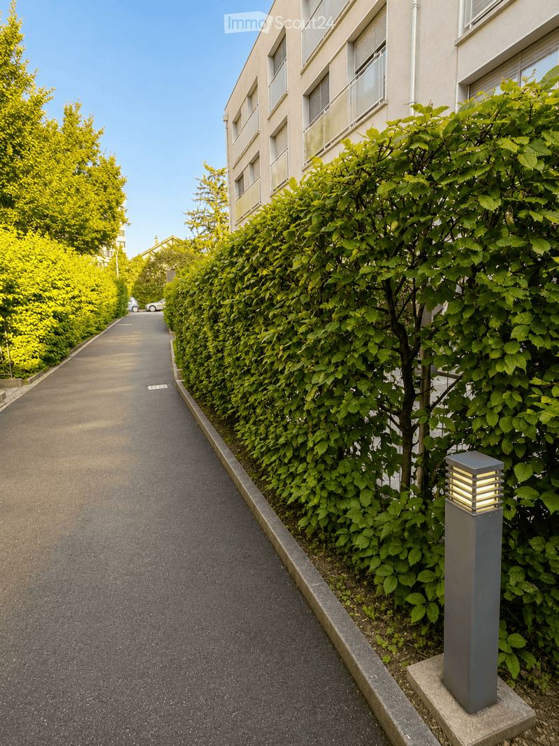 A quiet residential street lined with tall, neatly trimmed hedges and modern street lamps, with a multi-story building visible in the background.