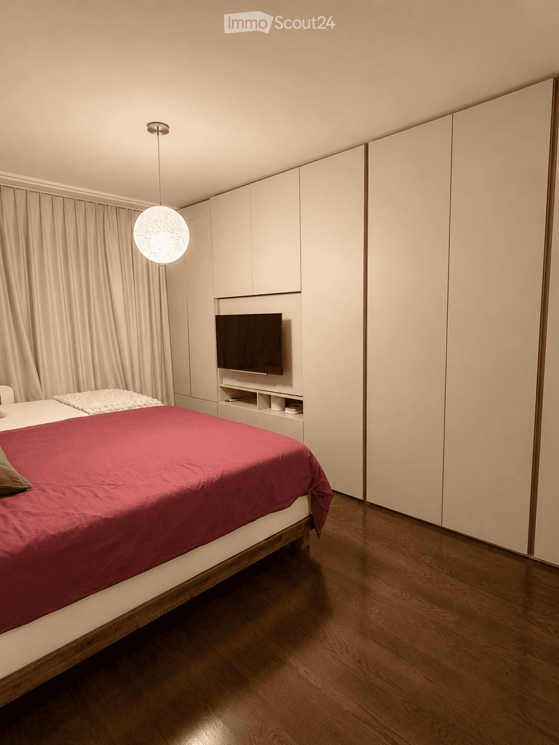 Modern bedroom with a large bed covered in a red duvet, a built-in wardrobe, a television mounted on the wall, and a pendant light fixture.