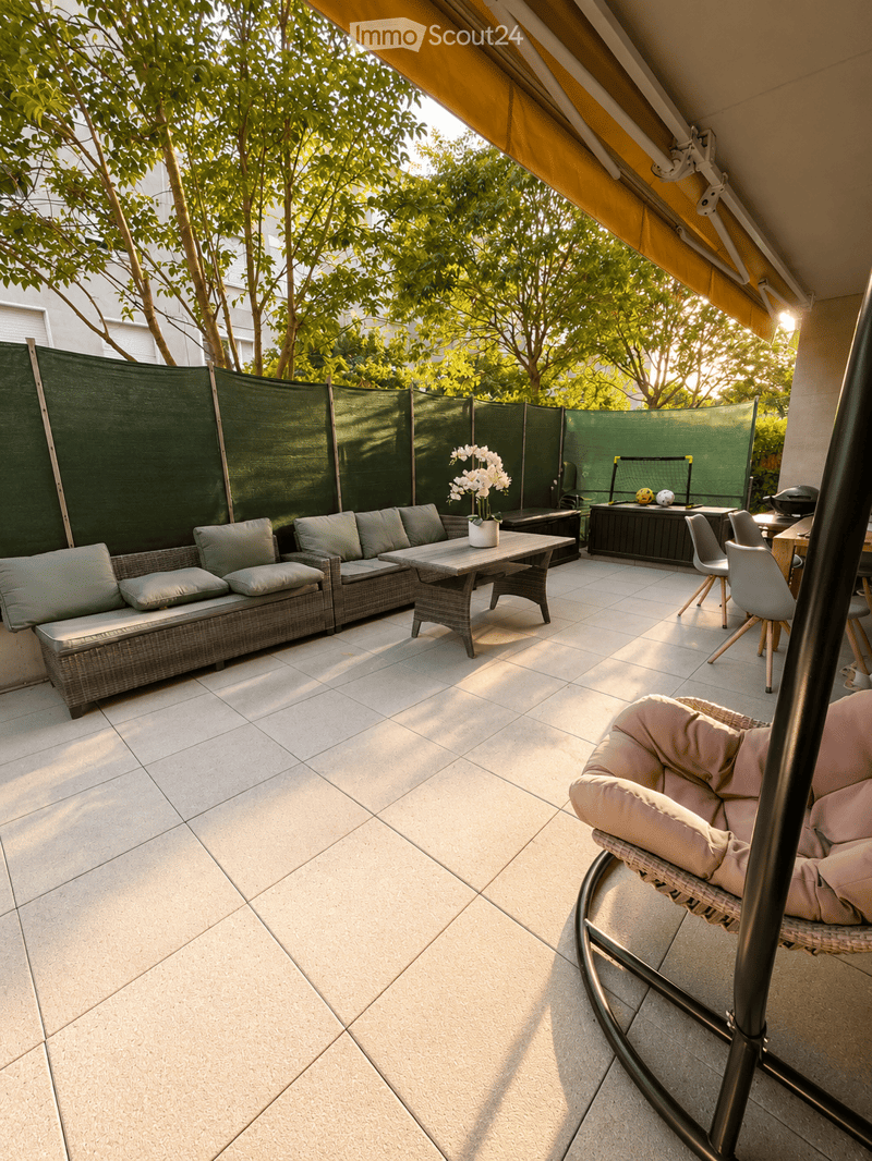 A well-maintained outdoor patio with a tiled floor, a wicker sofa, a coffee table, a hanging chair, and a small table with chairs. Green privacy screens and a yellow awning provide shade, with trees and a building visible in the background.